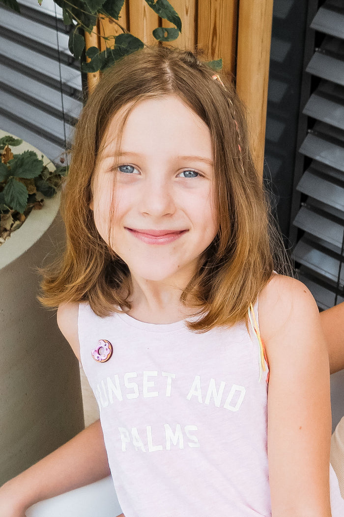 A young girl with shoulder-length light brown hair and blue eyes smiles at the camera. She wears a pale pink sleeveless top reading SUNSET AND PALMS, paired with a handcrafted Donut Pin. Plants and wooden details decorate the background.