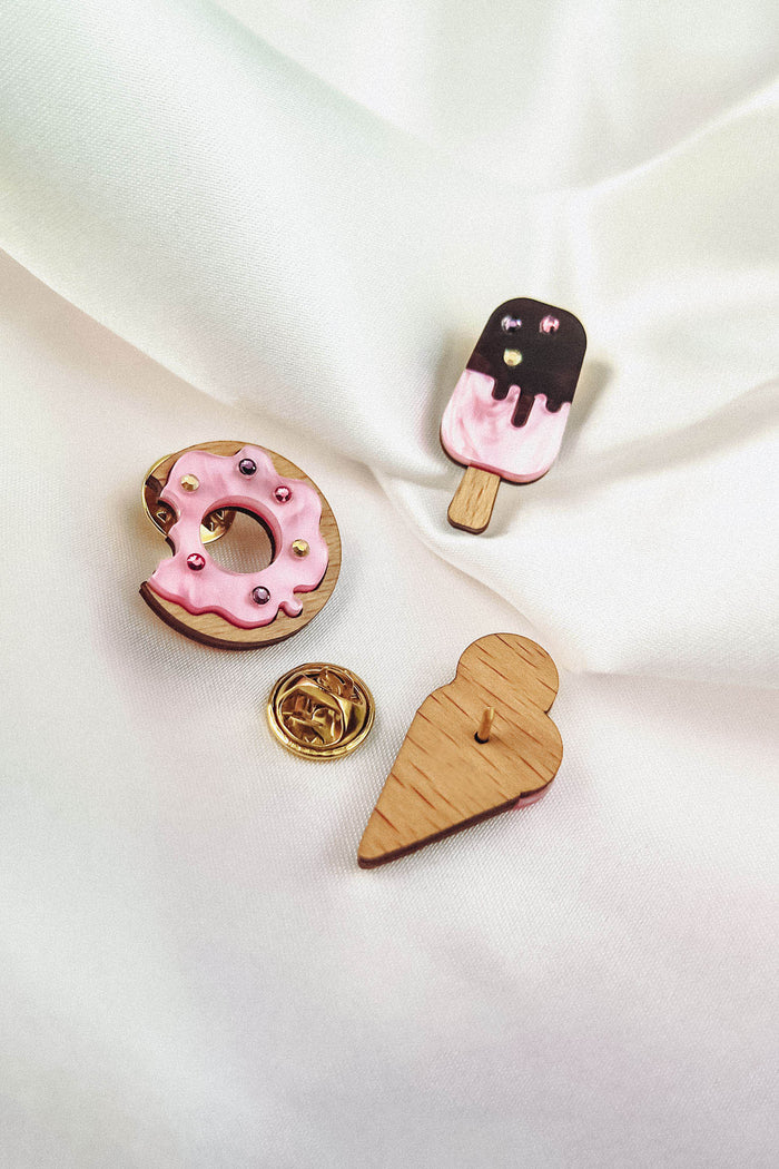 Three handcrafted wooden pins on white fabric: a pink frosted Donut Pin with sprinkles, a pink and brown ice cream pop adorned with colorful rhinestones, and a plain wooden heart. One gold pin backing is also visible.