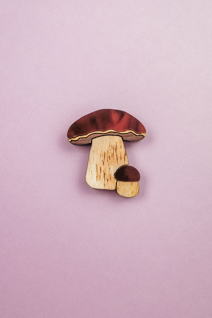A wooden King Bolete Mushroom Pin, featuring brown caps and light stems, showcases handcrafted quality and a nature-inspired design against a pastel pink background.