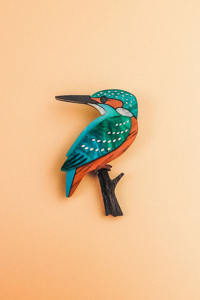 A colorful handcrafted Kingfisher brooch with teal, orange, and black details sits on a beige background, beautifully made from sustainable materials.