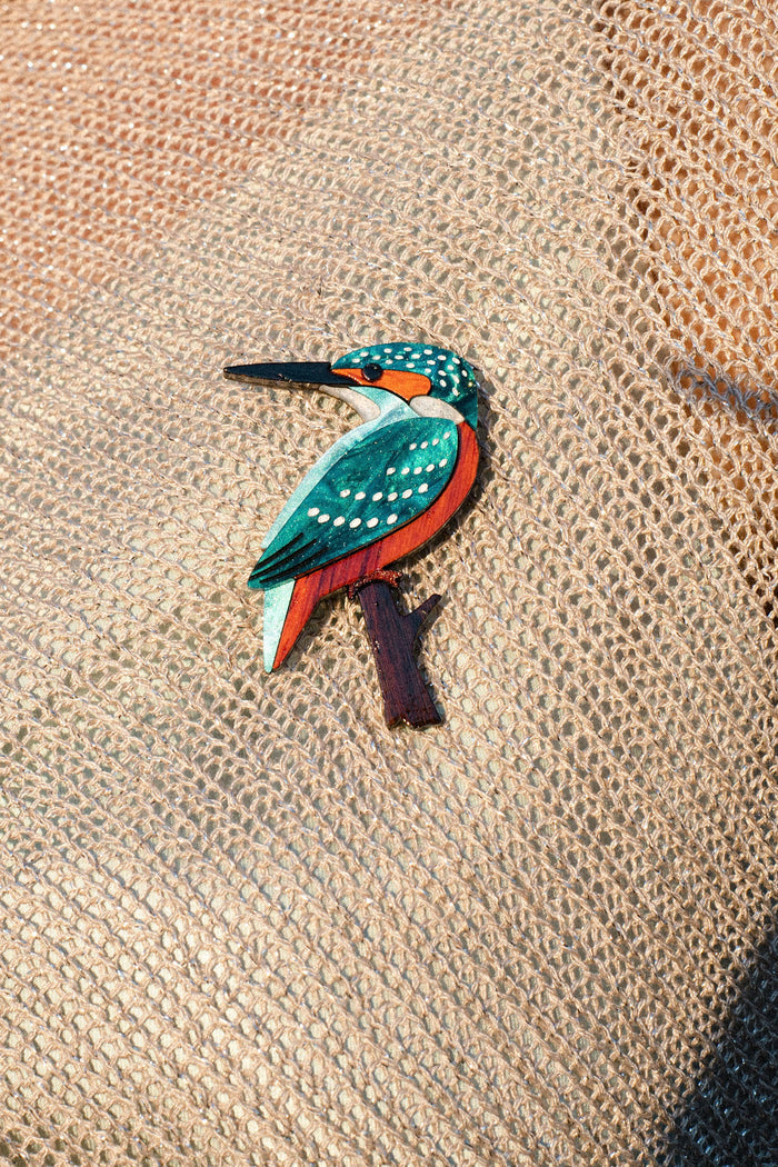 A handcrafted Kingfisher Brooch, painted on wood and made from sustainable materials, lies on textured brown burlap fabric under natural sunlight.