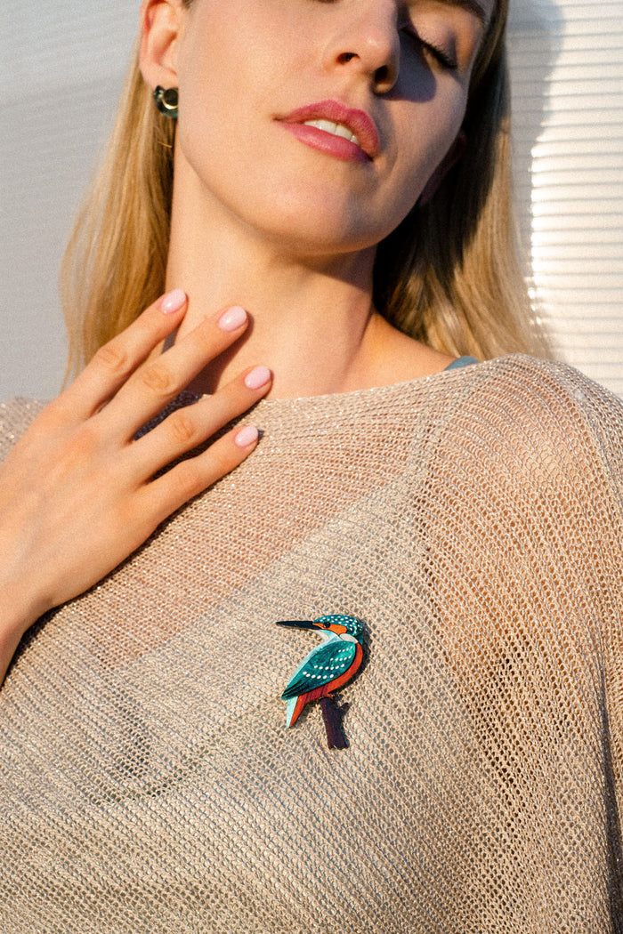 A woman wearing a sheer beige top with a colorful Kingfisher Brooch, handcrafted from sustainable materials, touches her chest lightly with one hand. Her face is relaxed, and she stands in soft sunlight.