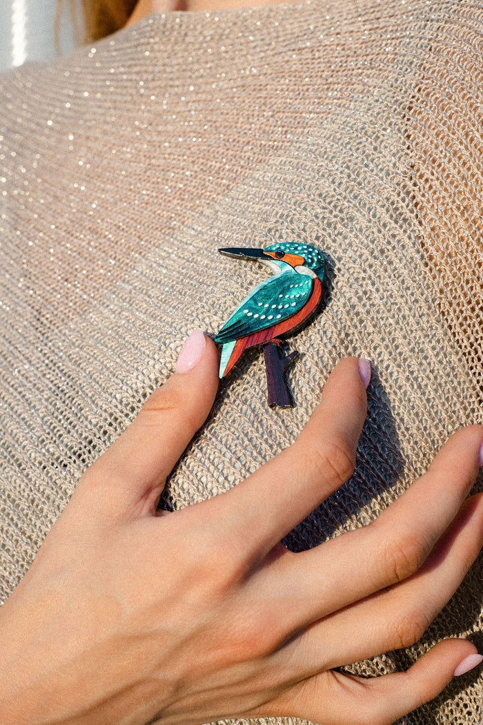 A hand with light pink nails touches a sparkling beige top adorned with a colorful kingfisher brooch, beautifully handcrafted from sustainable materials.