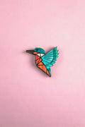 A colorful, geometric kingfisher pin brooch shaped like a bird with turquoise wings and head, and an orange body, handcrafted from recycled wood, set against a solid pink background.