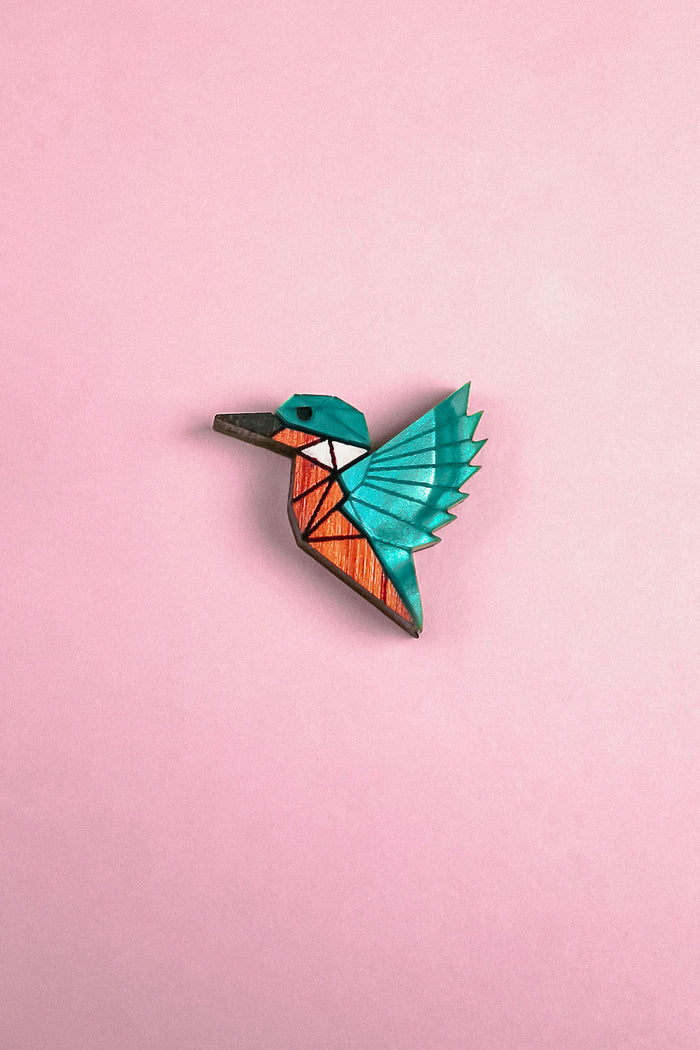 A colorful, geometric kingfisher pin brooch shaped like a bird with turquoise wings and head, and an orange body, handcrafted from recycled wood, set against a solid pink background.