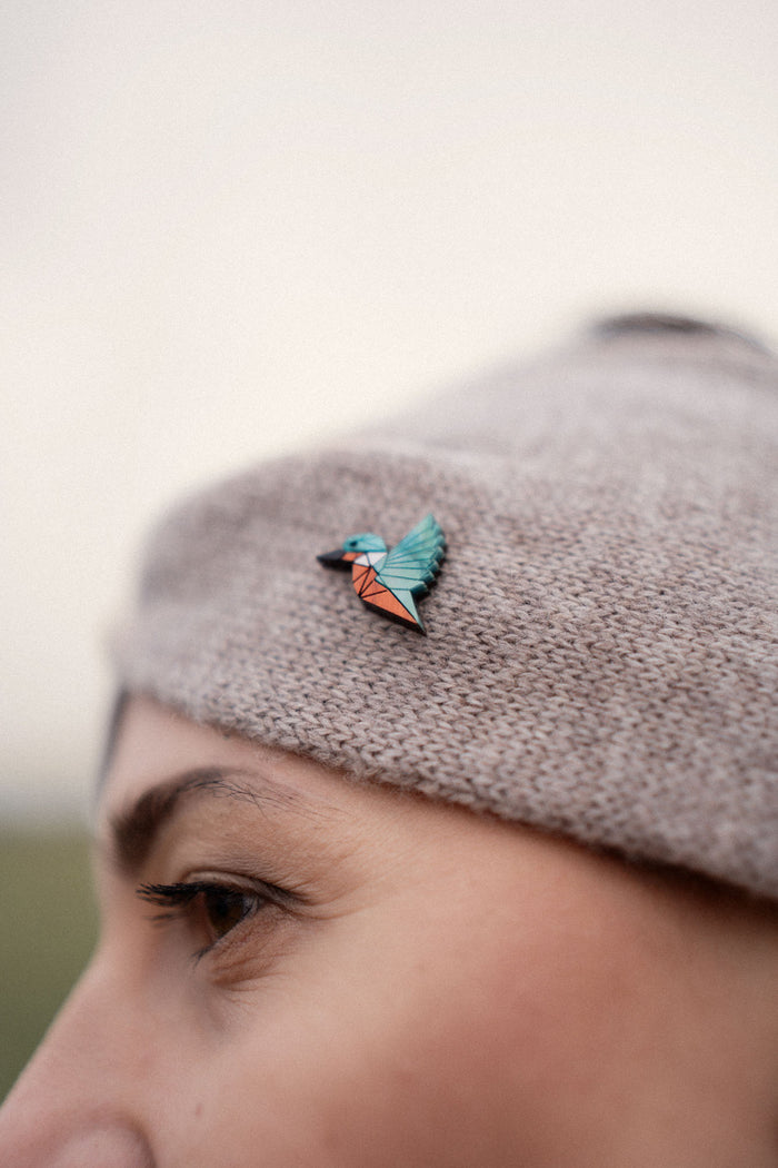 A close-up of a person wearing a knit beige hat decorated with a colorful kingfisher pin in shades of teal, blue, and orange. This eco-friendly accessory is perfect for bird lovers. The person's eyebrow and eye are partially visible.