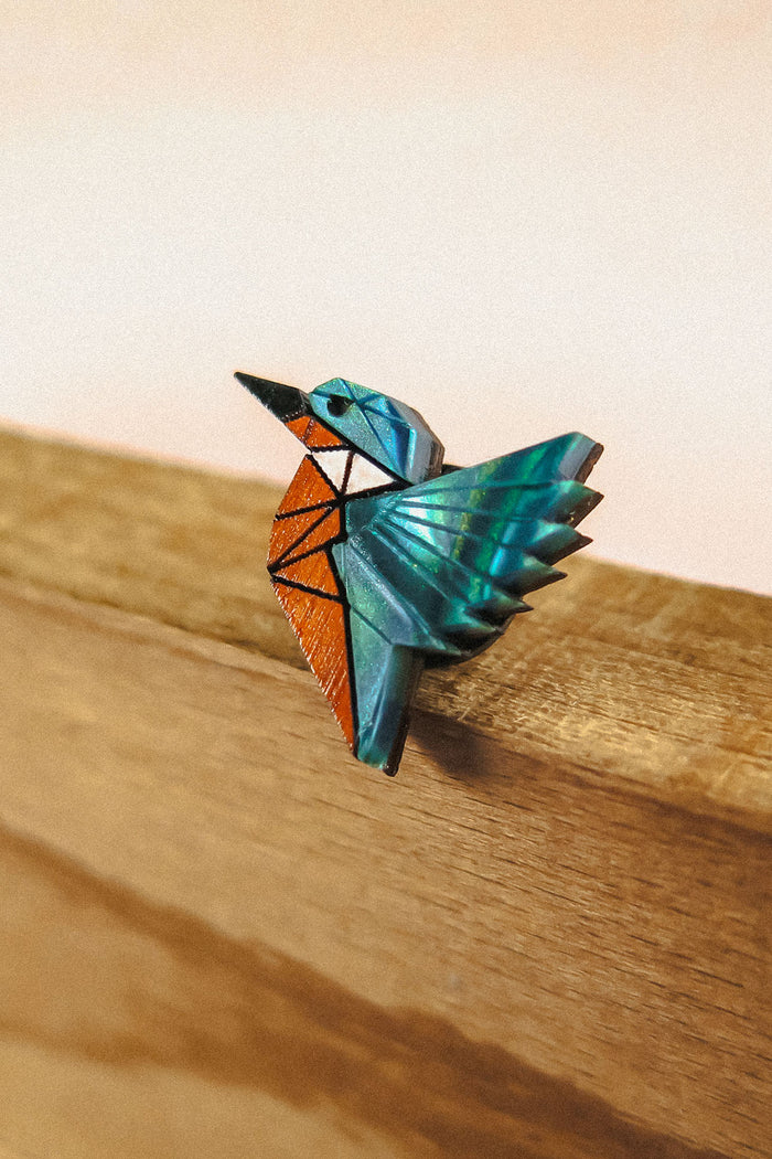 A colorful origami-style kingfisher pin brooch with blue, teal, and orange geometric sections, perched on the edge of recycled wood against a soft beige background—crafted from eco-friendly materials.