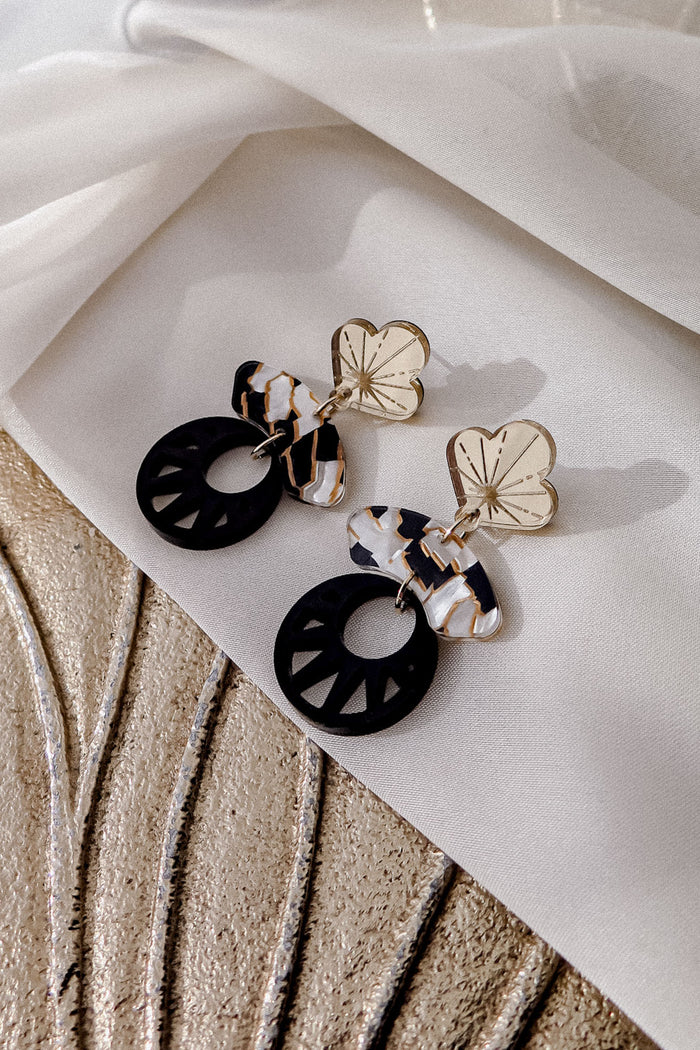 A pair of stylish Kintsugi Earrings featuring gold flower-shaped studs, marbled white and brown connectors, and large black circular pendants, all handcrafted and displayed on a textured beige fabric background.