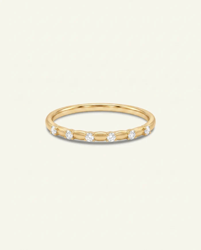 A delicate yellow gold eternity ring featuring five round-cut clear gemstones set in a row, presented on a clean, solid off-white background.