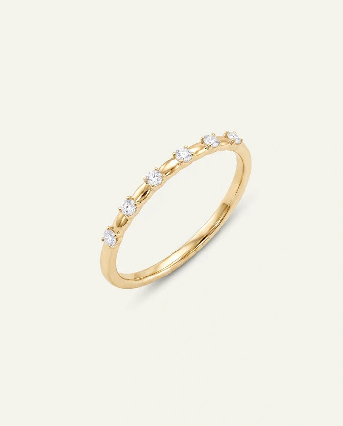 A delicate yellow gold ring featuring a row of small, round-cut clear gemstones, photographed on a clean, solid off-white background.