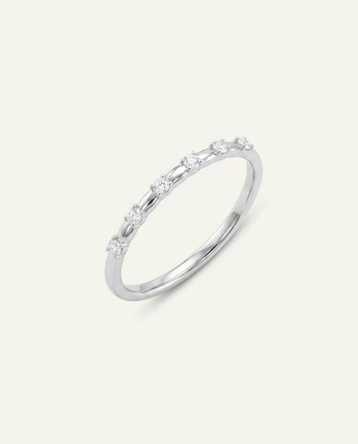 A delicate silver-toned or white gold ring featuring a row of small, round-cut clear gemstones set along the top of the band, presented on a light cream background.