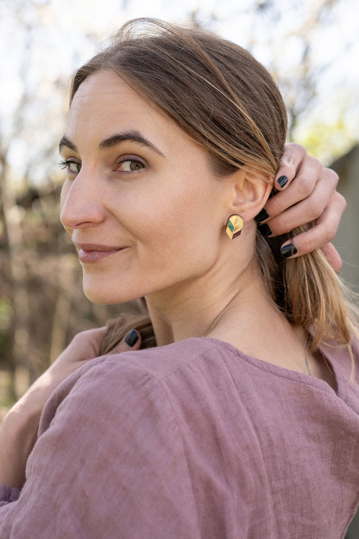 A woman with light brown hair in a low ponytail stands outdoors, wearing a mauve blouse and minimalist jewelry—gold leaf studs that catch the light. She looks over her shoulder at the camera, smiling softly, with one hand touching her hair.