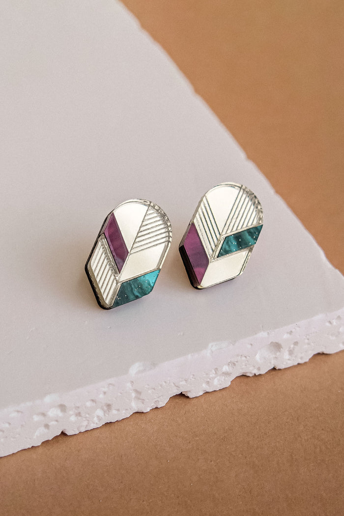 A pair of geometric stud earrings with silver outlines, featuring sections of teal, purple, and cream enamel, perfect for lovers of minimalist jewelry and displayed on a textured white and brown surface.