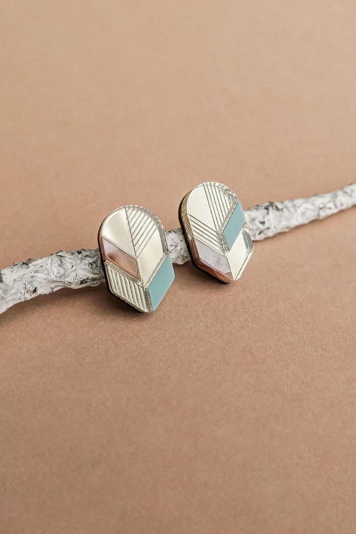 A pair of geometric Leaf Studs in gold, silver, and teal rest on a textured white branch against a light brown background—perfect for lovers of minimalist jewelry.