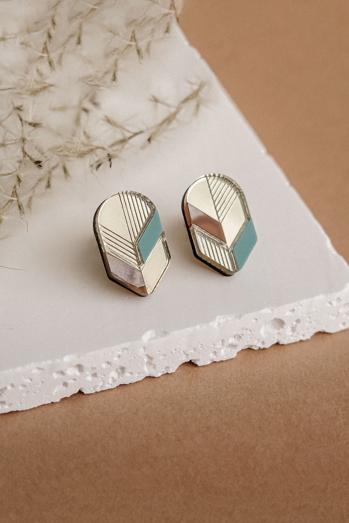 A pair of handmade geometric stud earrings with gold, pink, white, and teal sections, displayed on a white textured slab against a beige background with dried botanical accents—perfect for fans of minimalist jewelry.