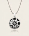 A silver compass pendant necklace on a matching chain, featuring a black compass rose design in the center with white lettering, surrounded by a beaded silver border, set against a light cream background.