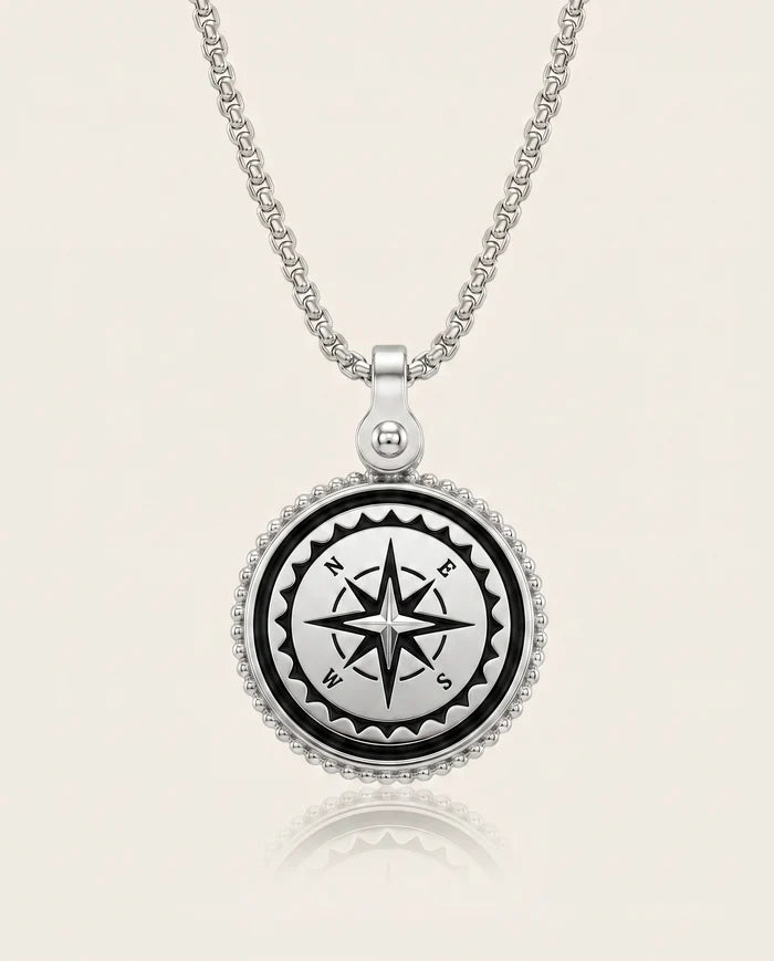 A silver compass pendant necklace on a matching chain, featuring a black compass rose design in the center with white lettering, surrounded by a beaded silver border, set against a light cream background.