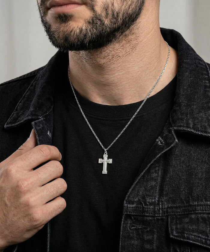 A close-up of a man wearing a silver cross pendant necklace over a black t-shirt and dark denim jacket.