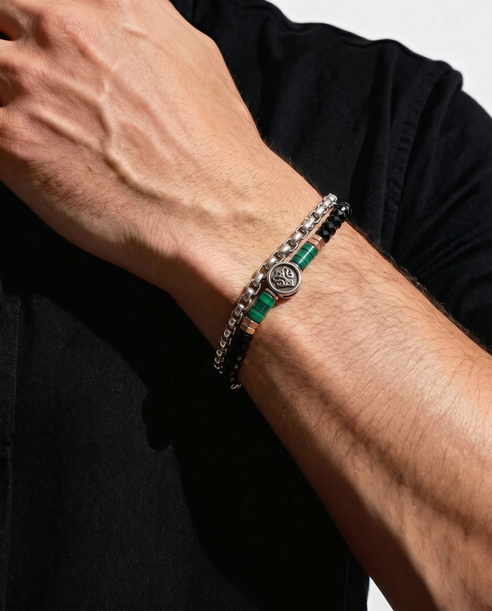 A close-up of a man's forearm wearing a stack of three bracelets. The bracelets include a silver chain with clear stones, a black beaded bracelet with green accents, and a silver chain with a circular medallion.