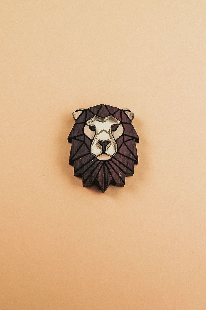 A handcrafted geometric Lion Pin brooch with a dark brown mane and light beige face, displayed on a plain light tan background.