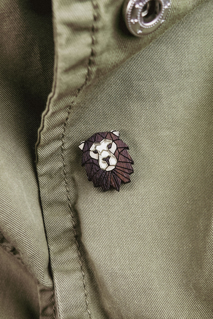 A close-up of an olive green jacket with a handcrafted geometric accessory—a Lion Pin—attached near a button.