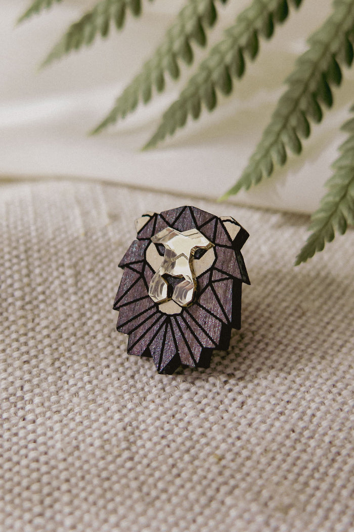 A handcrafted geometric lion pin with metallic gold and dark purple facets rests on textured beige fabric, complemented by green fern leaves in the background.