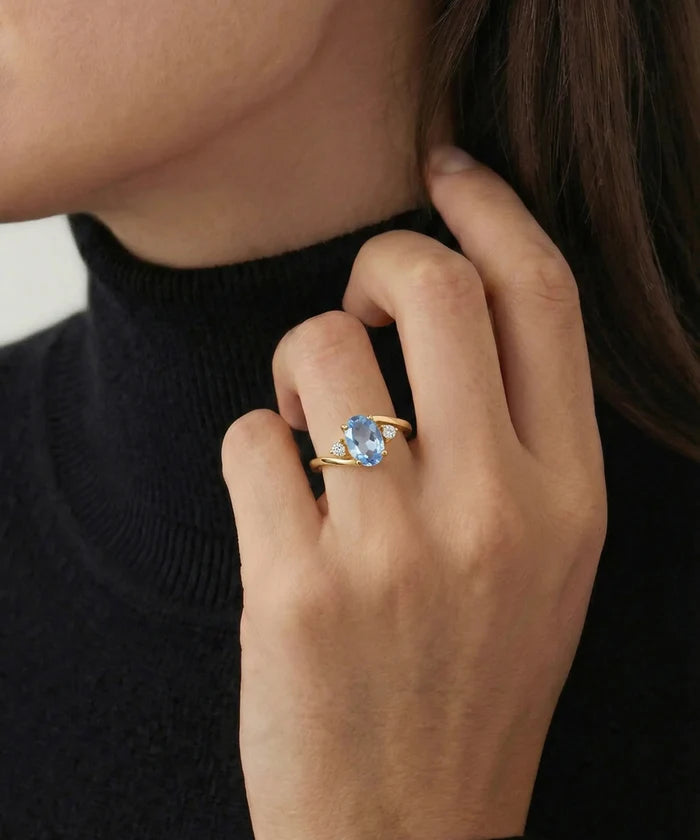 A close-up of a person wearing a gold ring with a light blue oval gemstone, shown on their finger against a black turtleneck sweater.