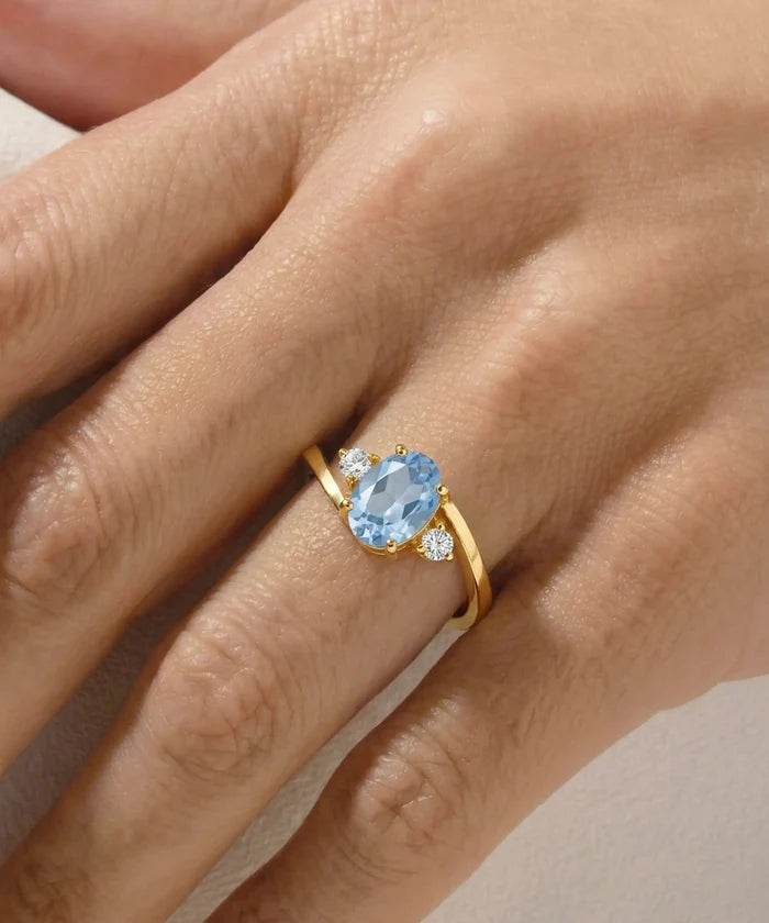 A gold ring with an oval light blue gemstone and small clear accent stones, shown worn on a person's finger.