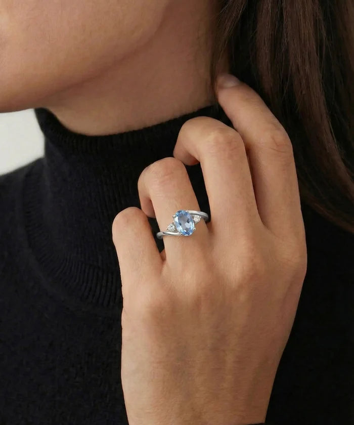 A close-up of a person wearing a silver ring with a light blue cushion-cut gemstone on their ring finger, paired with a black turtleneck sweater.