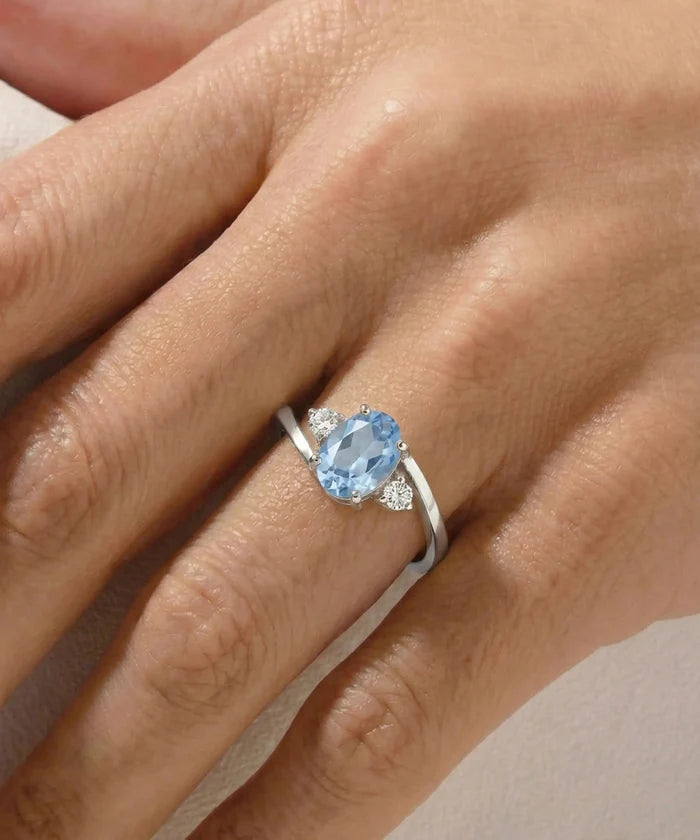 A silver ring featuring an oval-cut light blue gemstone and small clear accent stones, shown worn on a person's finger.
