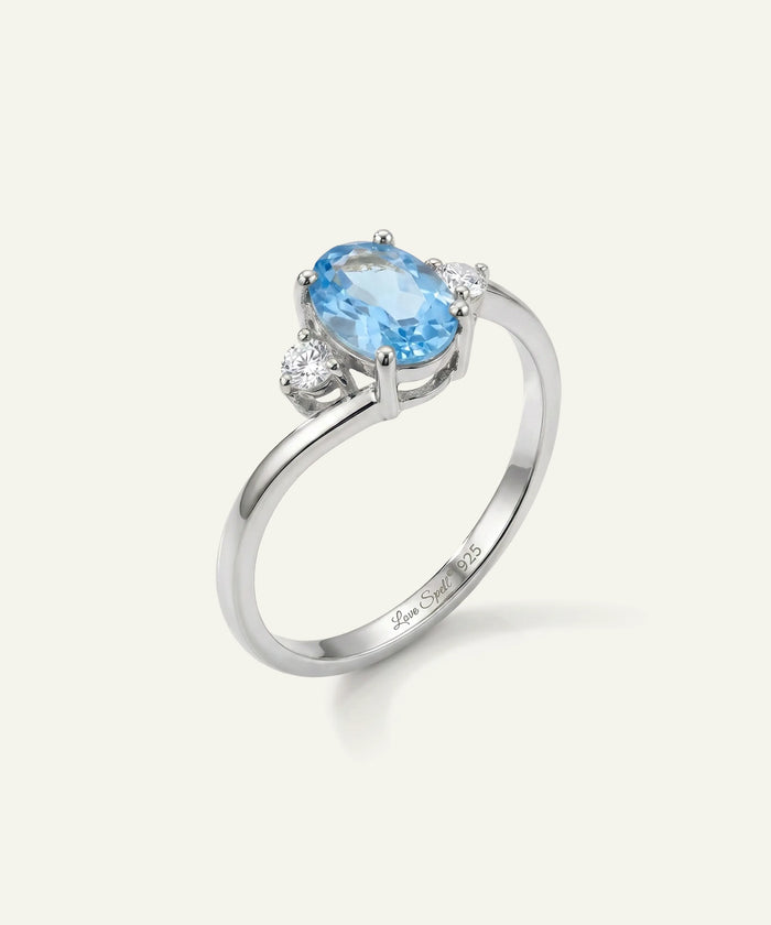 A silver ring featuring an oval-cut light blue gemstone in a prong setting, accented by two small round clear stones on either side, shown on a light cream background.