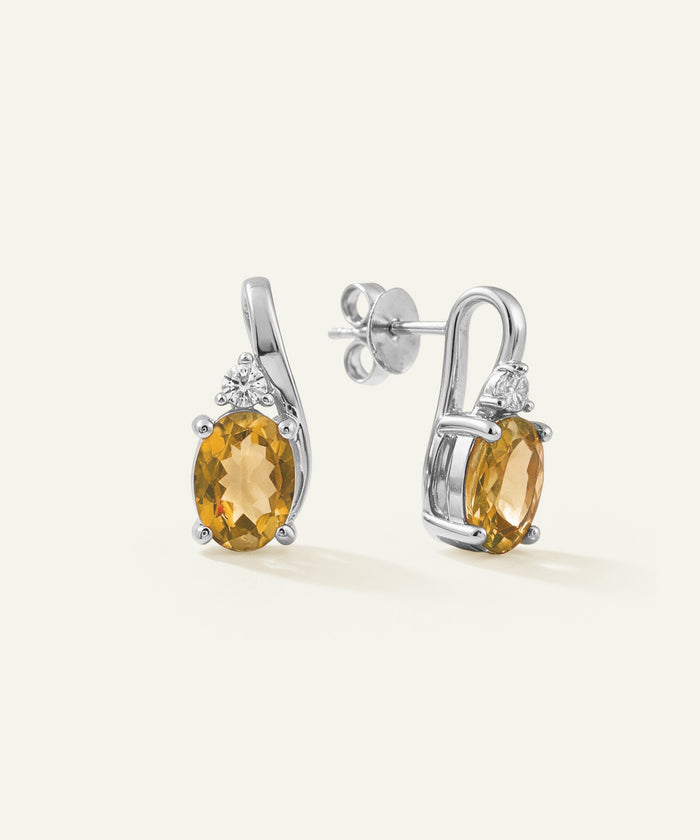 A pair of silver drop earrings featuring oval yellow-gold gemstones and small clear accent stones, shown on a clean off-white background.
