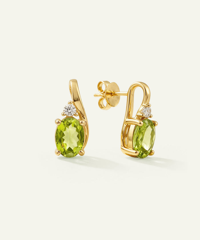 A pair of gold stud earrings featuring oval green peridot gemstones and small clear accent stones, shown on a clean off-white background.