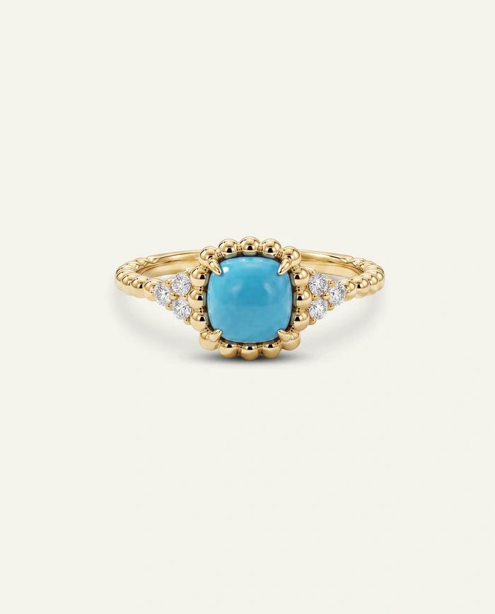 A gold ring featuring a cushion-cut turquoise center stone and small clear accent stones on a cream background.