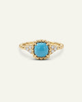 A gold ring featuring a cushion-cut turquoise center stone and small clear accent stones on a cream background.