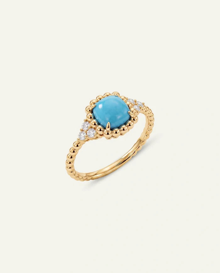 A gold ring featuring a central oval turquoise stone surrounded by small clear stones and a beaded gold band, set against a solid off-white background.
