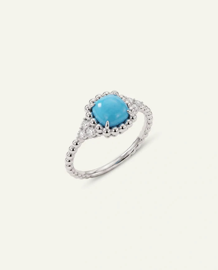 A silver ring featuring a cushion-cut turquoise center stone surrounded by a halo of small clear stones, with a beaded band, set against a light cream background.