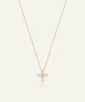 A delicate gold chain necklace with a small floral cross pendant set with clear gemstones, displayed on a light cream background with a soft reflection below.