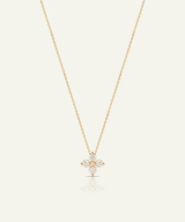 A delicate gold chain necklace with a small floral cross pendant set with clear gemstones, displayed on a light cream background with a soft reflection below.