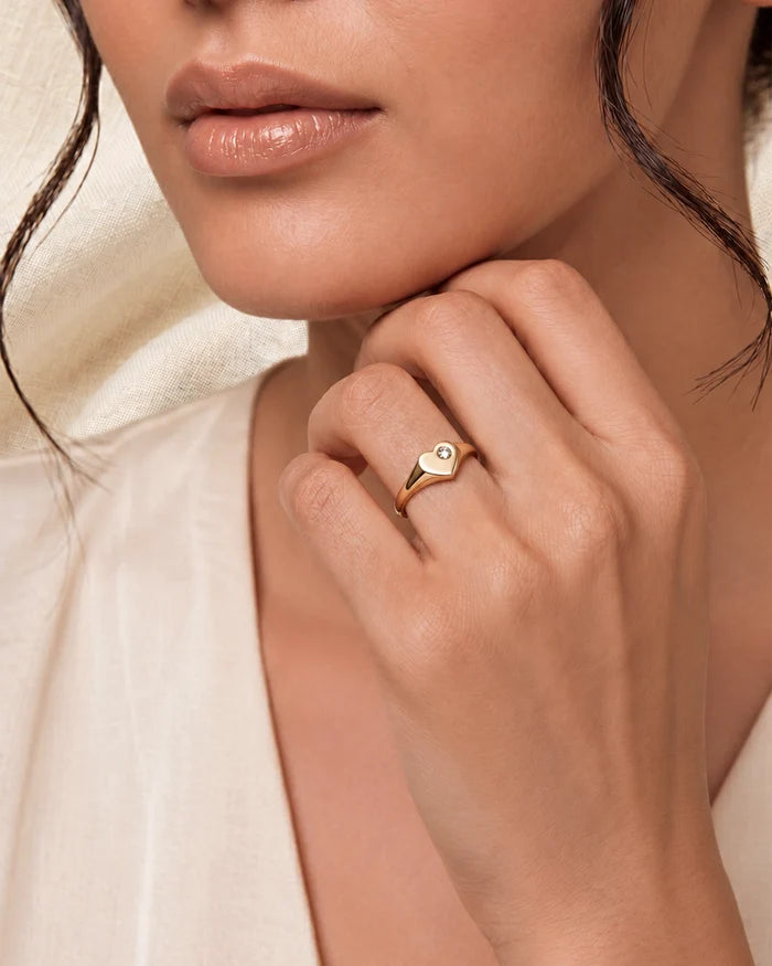 A close-up lifestyle shot of a model wearing a delicate gold ring with a small white enamel heart on a sliver band, styled with a cream-colored top.