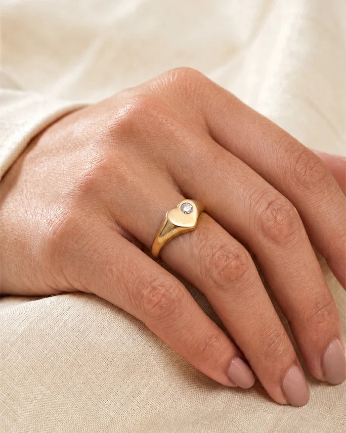 A gold signet-style ring with a small clear gemstone, worn on a person's ring finger against a soft beige fabric background.