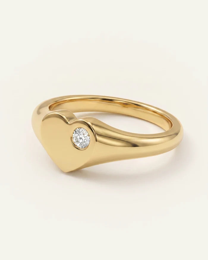A polished yellow gold heart-shaped ring with a single round-cut clear gemstone set on the front, photographed on a clean off-white background.