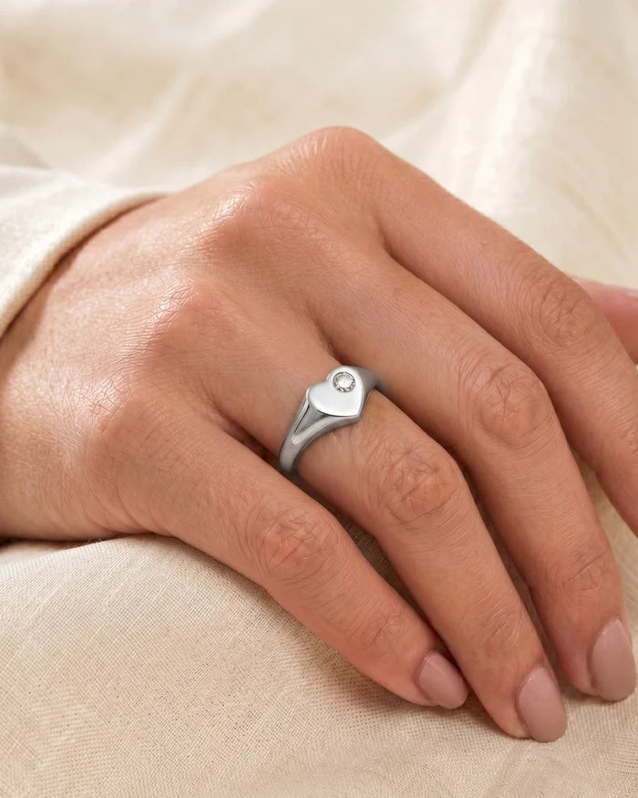 A silver signet-style ring with a small clear gemstone, worn on a person's finger against a soft beige fabric background.