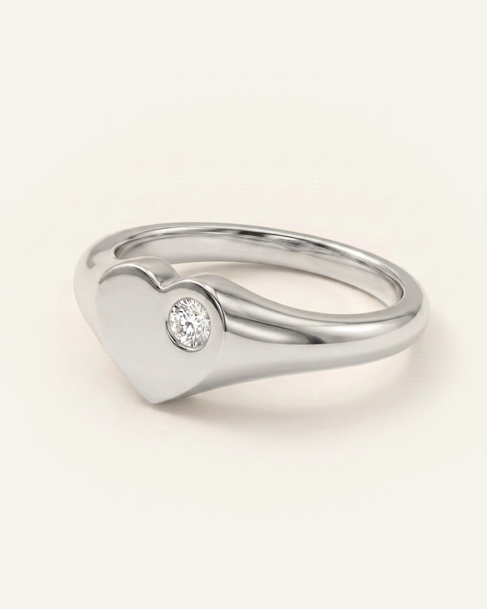 A polished silver heart-shaped ring with a small round clear gemstone on the front, set against a plain off-white background.