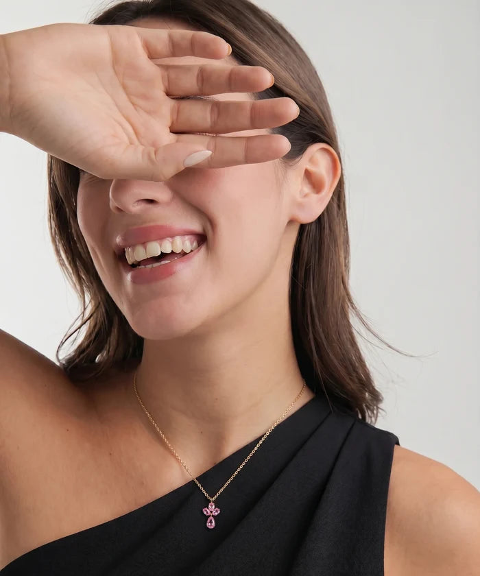 A close-up lifestyle shot of a woman wearing a delicate gold chain necklace with a pink bow pendant, with her hand covering part of her face against a light gray background.