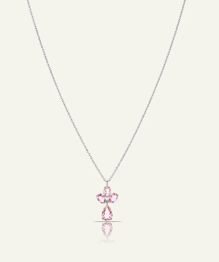 A delicate silver-toned chain necklace featuring a small cross-shaped pendant with pink gemstones and a teardrop pink stone hanging below, set against a light cream background with a soft reflection.