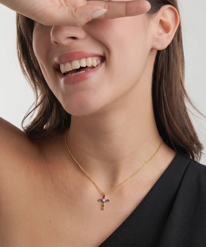 A close-up lifestyle shot of a woman wearing a delicate gold chain necklace with a small cross pendant adorned with colorful gemstones, set against a clean light gray background.