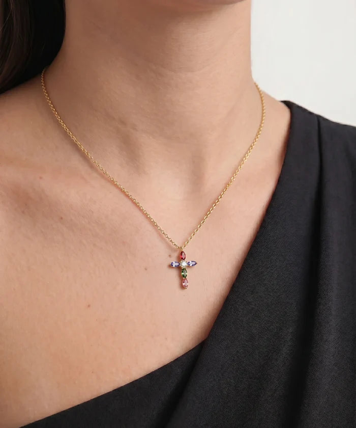 A close-up lifestyle shot of a woman wearing a delicate gold chain necklace with a small cross pendant adorned with colorful gemstones, set against a clean light gray background.
