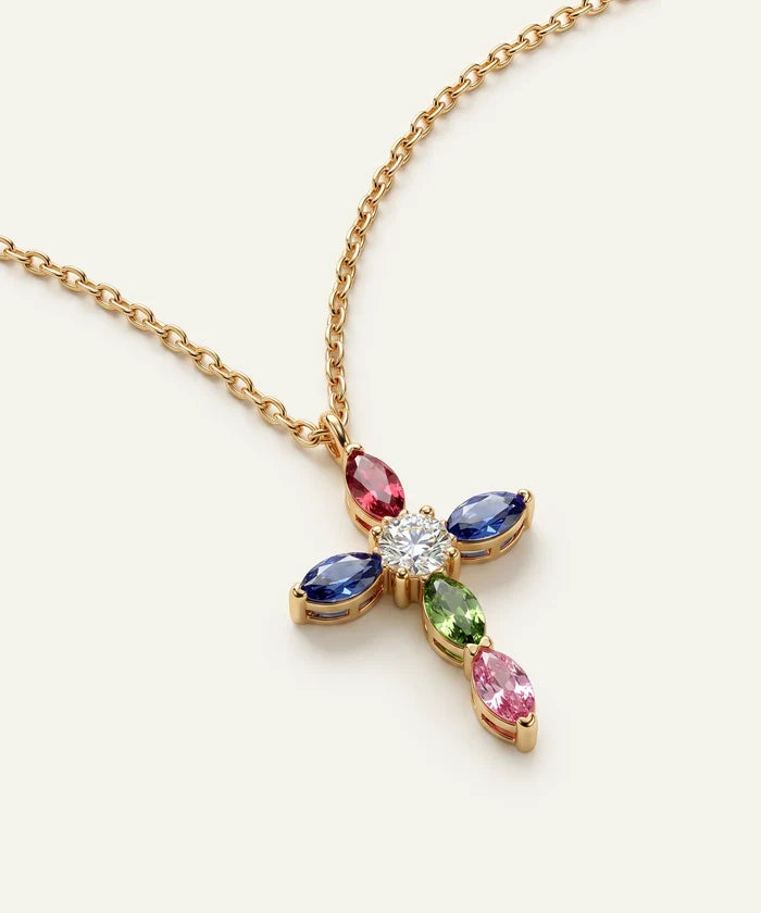 A gold cross pendant necklace featuring a central clear round gemstone surrounded by marquise-cut pink, blue, and green gemstones, suspended from a delicate gold chain on a light cream background.