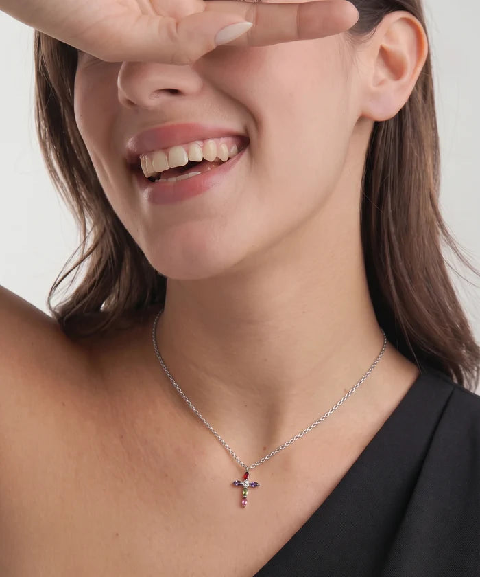 A close-up of a woman wearing a delicate silver cross pendant necklace with small red and blue gemstone accents, with her hand covering part of her face.